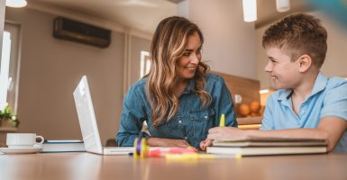 Photo of Mother helping her son with homework in the kitchen at home. Mature brown hair Mother looking after son doing homework on laptop. Beautiful business woman and her cute little son are drawing and smiling while sitting. Mother helping her son to prepare for written assignment.